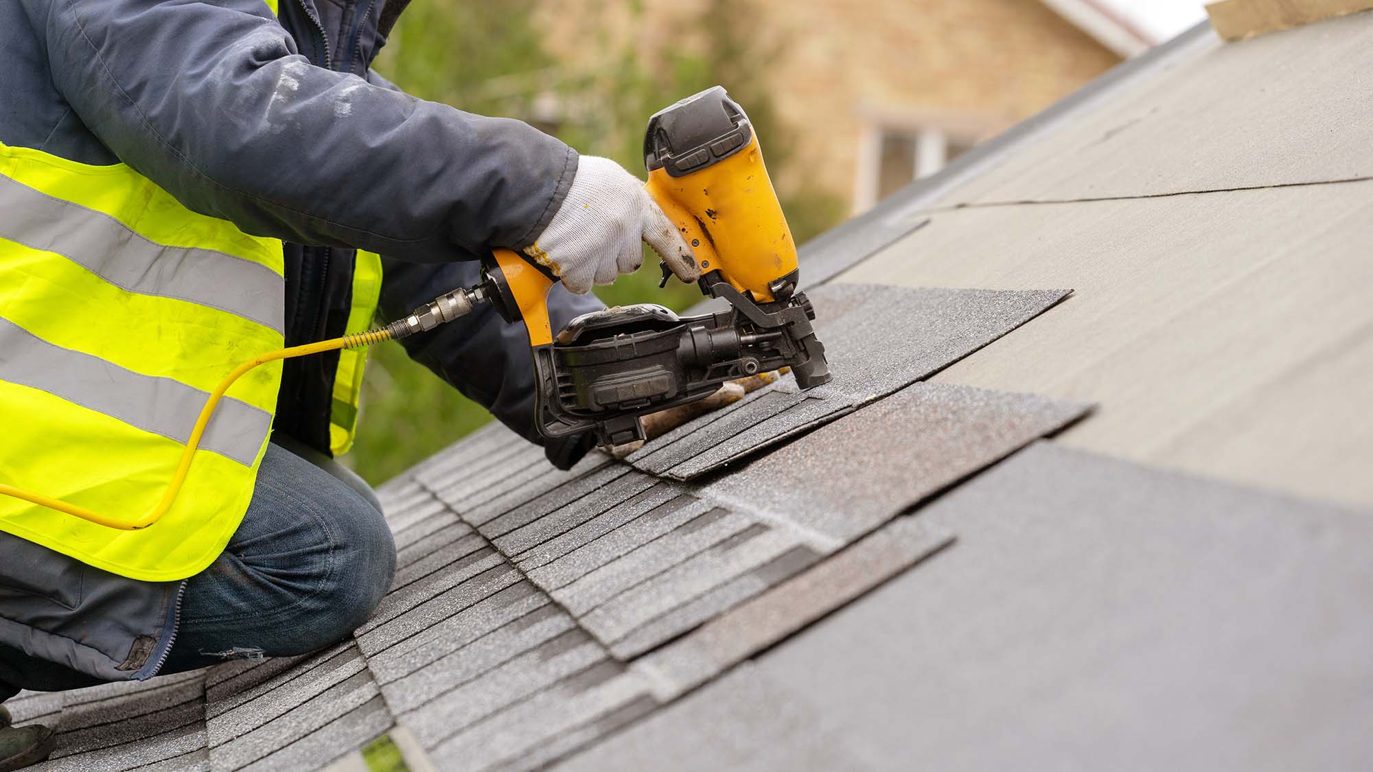 Roofer worker in special protective work wear and gloves, using air or pneumatic nail gun and installing asphalt or bitumen shingle on top of the new roof under construction residential building